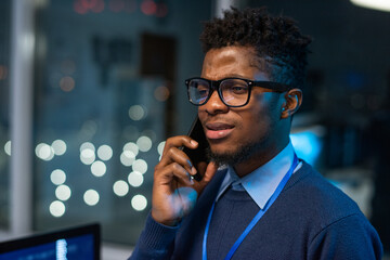 Poster - Young businessman in eyeglasses speaking on smartphone in front of camera against office environment