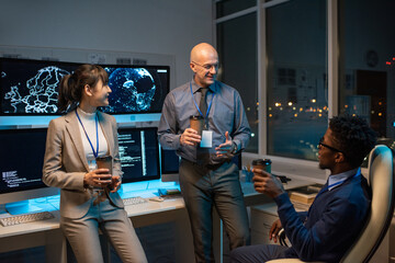 Poster - Three happy intercultural colleagues with drinks chatting at break by workplace with computer monitors