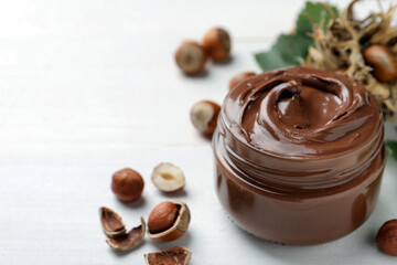 Poster - Glass jar with tasty chocolate spread and hazelnuts on white wooden table, space for text