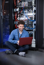 Smiling IT Engineer Using Laptop While Sitting In Front Of Rack In Server Room