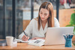 © Daenin - Female student taking notes from a book computer and smartphone at the table An Asian woman sits at an assignment desk in a college library.