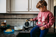 Young Girl Sitting On Counter In Modern Kitchen Pouring Waffle Batter