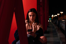Young Woman Is Sitting And Is Looking At Her Telephone On A Red Bridge