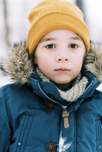 Little Boy In Thick Winter Coat And Yellow Knit Hat Looking Serious