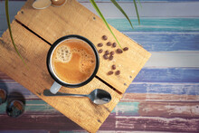 A White Coffee Cup With Cream Coffee Stands On A Colored Wooden Wooden Surface. Coffee Beans Are Next To It