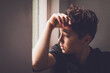 © Cavan Images - Boy leaning against the wall looking outside a window.