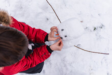 Overhead View Of Child In Red Coat Building A Snowman On Winter Day.