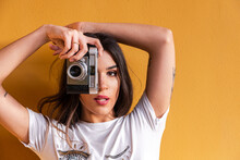 Portrait Of Spanish Brunette Girl Taking A Photo With A Vintage Camera On Yellow Background Wall.