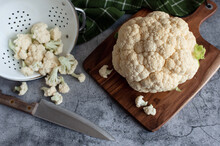 Cauliflower And Florets On Cutting Board And Colander On Gray Counter.