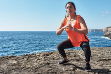 Latin Woman, Middle-aged, With Orange Top, Black Leggings, Training, Doing Physical Exercises, Squats, Burning Calories, Keeping Fit, Outdoors By The Sea, Blue Sky, Day, Sunny Winter, Bluetoo