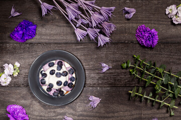 Blueberry Graham Cheesecake topped with fresh berries, on a dark wood background with purple flowers and twigs. Flat lay, top view.