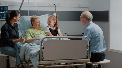 kid hugging sick grandma at family visit in hospital ward. mother, little girl and old man talking t