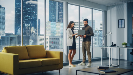 Successful Corporate CEO and Investment Manager Talking, Using Laptop Computer while Standing in Big City Office. Two Successful Diverse People Brainstorm Their e-Commerce Software Investment Strategy