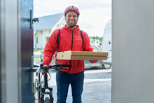 Smiling Deliveryman Holding Pizza Box While Standing With Bicycle At Doorway