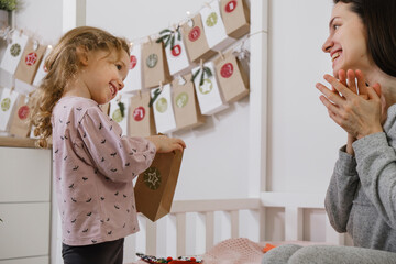 Mother with child opening Christmas advent calendar tasks and gifts. Toddler girl excited about festive surprise in craft bag at home. Family celebration and preparation for New Year