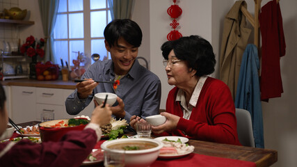 smiling asian adult son giving food to his senior mother as they are having reunion dinner together on Chinese new year's eve in the dining room at home