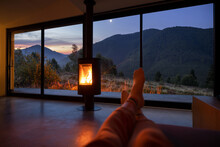 Woman Resting Near Fireplace At Modern House With Great View On Mountains At Sunset. Women's Legs On Background Of Landscape