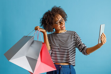 Wall Mural - Cheerful multiracial lady holding purchases and smiling while taking picture with smartphone. Isolated on blue background