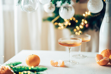 Wall Mural - Two glasses of champagne and tangerines on a table against blurred christmas background with christmas lights. New year celebration party