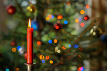 A Red Candle Against An Out-of-focus Christmas Tree With Lights On It. 