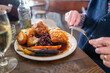 The golden thick crackling on top of roast pork belly on a white plate with roast potatoes and Yorkshire pudding with vegetables in a restaurant. A mans hands can be seen holding a knife and fork.