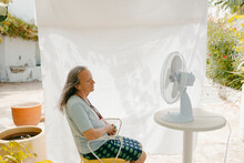 Mature Woman Sitting In Front Of Electric Fan Outdoors