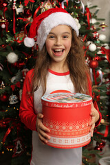 Wall Mural - a beautiful happy girl in a Santa Claus hat is standing at the Christmas tree with a gift box in her hands