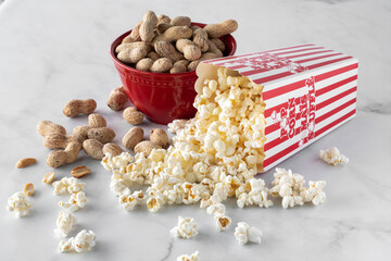 Close up of a container of popcorn tipped over with a bowl of peanuts in behind.