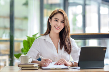 Wall Mural - Successful smiling beautiful young Asian businesswoman sitting with laptop and computer while doing some paperwork at the office. Looking at camera.