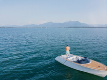 Germany, Bavaria, Shirtless Man Standing On Motorboat Floating In Chiemsee Lake Admiring View Of Distant Chiemgau Alps