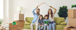 Portrait of happy family with kids sitting at home on sofa holding cardboard roof over their heads. Young family feels happy and protected in their new apartment. Insurance concept. Banner.