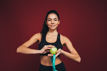 Smiling Female Athlete Holding Apple And Measuring Tape In Front Of Maroon Background