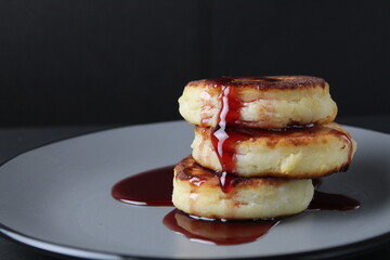 Canvas Print - food breakfast cheesecakes curd pancakes three pieces lie stack of slides on top of each other and pour jam sauce cherry red on a gray plate on a black background