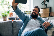 Smiling Man Taking Selfie Through Smart Phone While Sitting On Sofa At Home