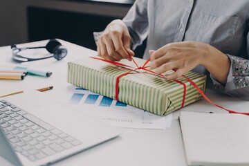 Cropped image of young businesswoman packing and decorating with ribbon Xmas gift at work