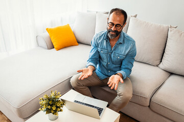 Wall Mural - Happy millennial mature man in glasses wearing headphones, enjoying watching educational webinar on laptop. Smiling mature mixed race businessman holding video call with clients partners.