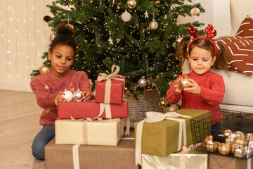 Two sisters in an interracial family unpacks gift boxes at home by the Christmas tree.The girls are dressed in red sweaters and jeans.New Year and Christmas concept.Selective focus.