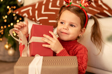 Little caucasian girl unpacks gift boxes at home by the Christmas tree.The girl is wearing a red sweater and jeans.Girl has a festive headband with deer horns on her head.New Year,Christmas concept.