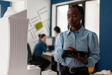 Poster - African america architect looking at scale foam model of modern building while holding tablet with blueprints. Architectural engineer focused on the design of 3d skyscraper maquette.