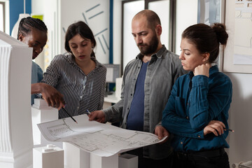 Poster - Team of architects analyzing blueprints for building project standing in front of table with real estate maquette. Professional engineers reading construction plans for urban development.