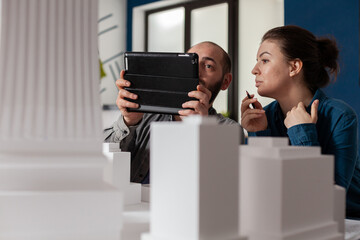 Poster - Project engineers working together looking at tablet with construction plans sitting at desk in modern architectural office. Two architects doing teamwork next to table with buildings maquette.