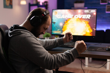 Wall Mural - Disappointed man losing video games on computer. Adult clenching fists and playing games with headphones and joystick, feeling sad about lost game. Player using modern equipment.