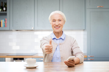 Canvas Print - technology, gesture and people concept - happy senior woman with smartphone and coffee sitting at table and showing thumbs up at home over kitchen background