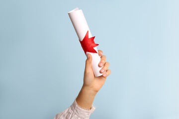Wall Mural - Student holding rolled diploma with red ribbon on light blue background, closeup
