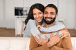 © Vadim Pastuh - Happy lovely young Indian couple together at home, ethnic young wife hugging from behind her husband, sitting and resting on sofa in modern apartment, portrait of romantic multiracial couple in love