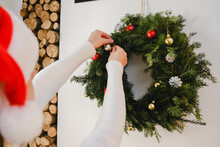 Selective Focus Of Young Woman Wearing Sweater And Santa Red Hat Decorating Christmas Wreath With Toys And Balls At Home, Preparing Home For New Year Celebration, Enjoying Holiday, Festive Interior