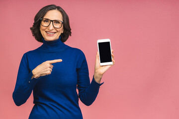 Happy smiling happy senior mature woman in casual showing blank smartphone screen while looking at the camera isolated over pink background. Using phone.