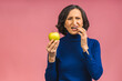 © denis_vermenko - Senior aged mature woman touching mouth with hand with painful expression because of toothache or dental illness on teeth holding green apple. Dentist concept isolated over pink background.