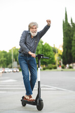 Happy Mature Man With Hand Raised Riding Electric Push Scooter On Street