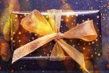 Wall Mural - Holiday gift box or present with ribbon, golden confetti and gold baubles on black background. Magic christmas greeting card. Christmas Decoration. Toned image with bokeh. Mock up. Banner.